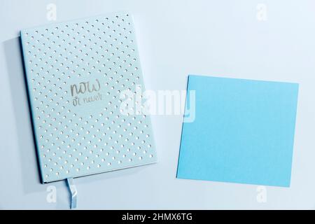Blue notebook in a white dot with text - now or never and dark blue paper on the surface of the table. Place for your text. Concept Stock Photo
