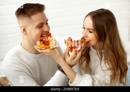 Enjoy pizza.Young woman and her lover are happy, having lunch together and jokingly feeding each other, they are sitting against white wall. Date at Stock Photo