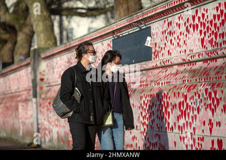 Two asian tourists with mask looking at the National COVID Memorial Wall painted with red hearts on beautiful sunny day . Westminster, London , UK Stock Photo