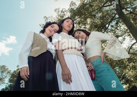 low angle view of three retro styled young girls standing in a plaza looking at the camera with the sun and trees behind them. vintage and lifestyle c Stock Photo