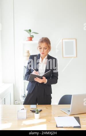 Nice businesswoman having been employed to job as executive standing near desktop with laptop noting down important information Stock Photo
