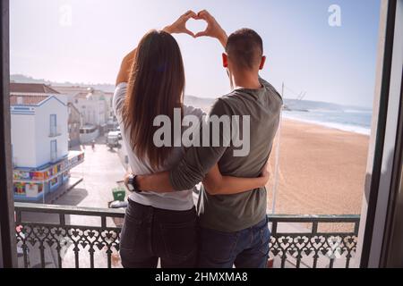 Young couple standing on the balcony and making heart shape with their hands. Couple in love hugging and spending valentine's day together Stock Photo