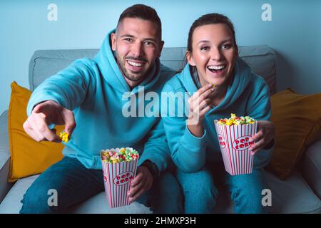 Young happy couple with popcorn sitting on sofa and watching funny comedy movie. Couple enjoying time together at home Stock Photo