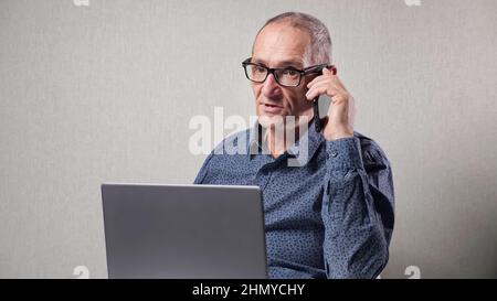 Elderly man talks on mobile phone working on laptop at table Stock Photo