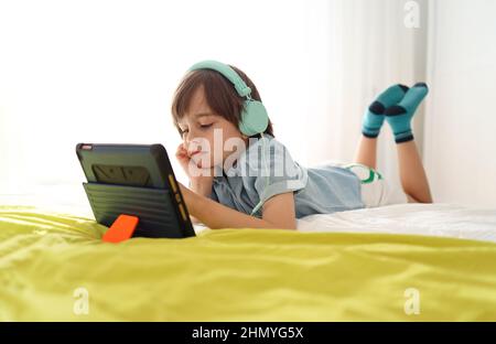 Bored little boy looking at tablet with sad face lying down on sofa looking deep in thought. Child using electronic devices while slouching on bed at Stock Photo