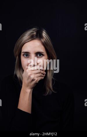 Threatened woman with hand on mouth making her be silent in black blouse on black background. Victim of physical and psychological abuse. Gaslighting Stock Photo