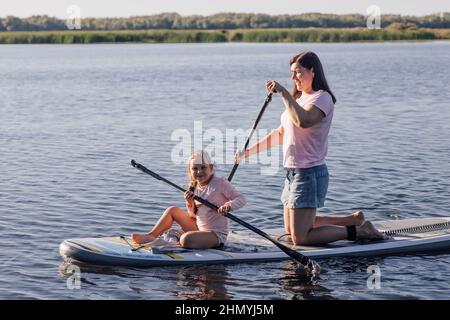 Middle aged mother teaching little female kid to do puddle boarding on lake with green trees and reeds in background. Active lifestyle. Teaching Stock Photo