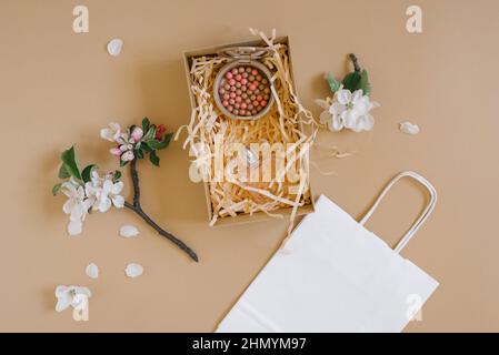 An open gift box with peach-colored paper shavings, which contains nail polish and blush in balls, a white paper bag and apple flowers on a beige back Stock Photo