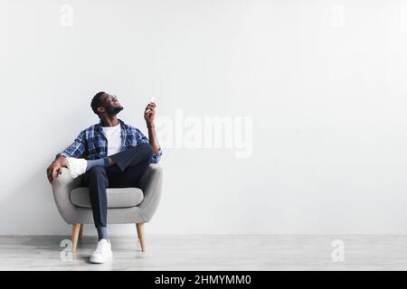 Split system. Young black man with remote turning on air conditioner, sitting in armchair against white wall, mockup Stock Photo