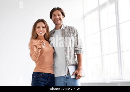 Satisfied happy european young couple in empty room with panoramic window, show keys to their own apartment Stock Photo
