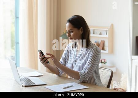Happy pretty young woman using cellphone, distracted from laptop work. Stock Photo
