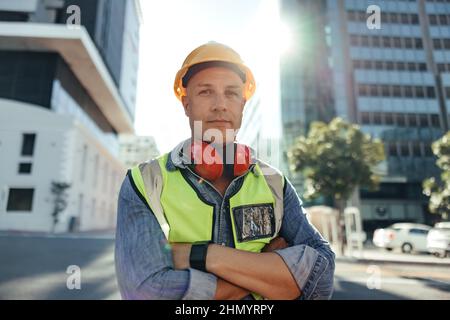 Confident construction worker looking at the camera outdoors. Mid-adult civil engineer standing with his arms crossed in the city. Blue collar worker Stock Photo