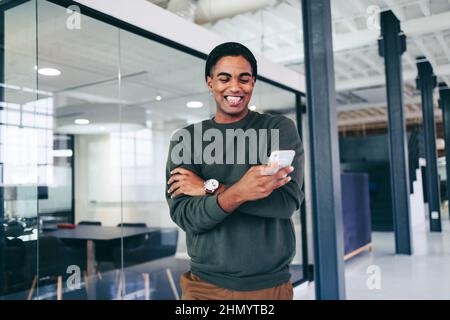 Good news in the office. Happy young businessman smiling cheerfully while reading a text message in a modern office. Successful businessman holding a Stock Photo