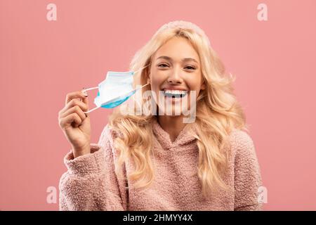 Coronavirus is over. Happy young caucasian lady taking off her face mask, standing on pink studio background Stock Photo