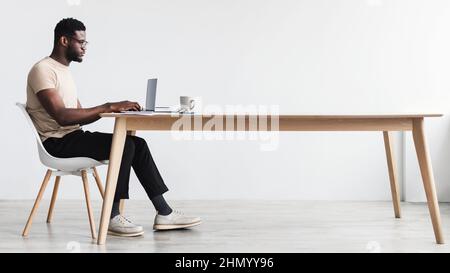 Side view of focused young black man using laptop, sitting at desk, working or studying online from home office Stock Photo