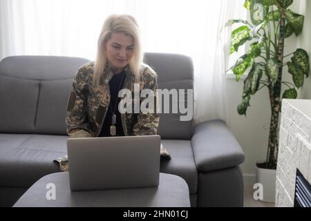Portrait of woman soldier wearing camouflage uniform and posing at home, sitting in front of laptop computer Stock Photo