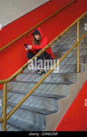 Young woman checking email on smartphone resting on stairs outdoors. Online message on cellphone Red colors. Stock Photo