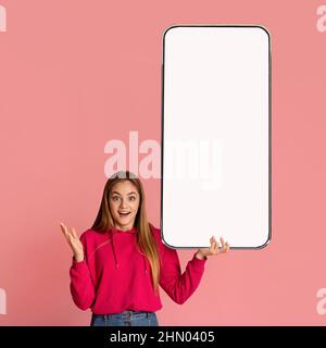 Check out this new mobile app. Excited woman presenting smartphone with white empty screen over pink background Stock Photo