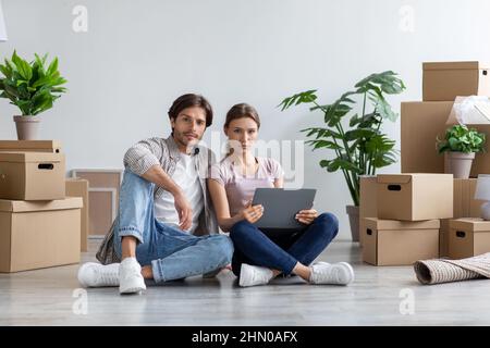 Serious european millennial lady and guy in casual look at tablet sit on floor with cardboard boxes Stock Photo