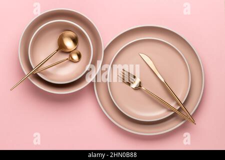 Dishes and utensils for serving and eating meals. Beige round rimmed plates and golden cutlery on a pink background, top view. Modern craft ceramic cr Stock Photo