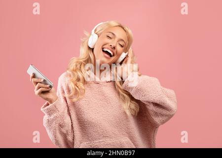 Overjoyed happy lady with headphones and smartphone listening to music and dancing over pink background Stock Photo