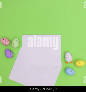 Paper card note in envelope and colorful vibrant eggs around on bright green background. Flat lay Stock Photo