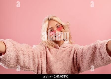Happy blogger lady making selfie, wearing colorful glasses and hoodie, posing and smiling to camera, pink background Stock Photo