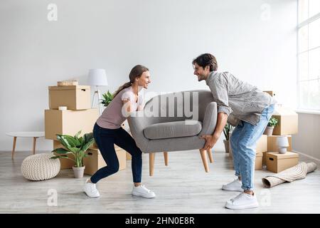 Smiling caucasian millennial wife and husband carry armchair in room with cardboard boxes Stock Photo