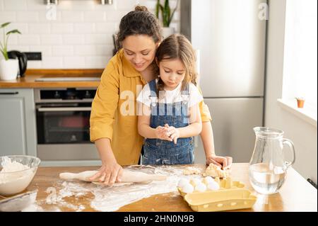 Middle-aged mother and cute toddler daughter cooking together, kneads the dough, using rolling pin, mom and kids spends leisure at home, baking cookies, enjoying weekend activity Stock Photo