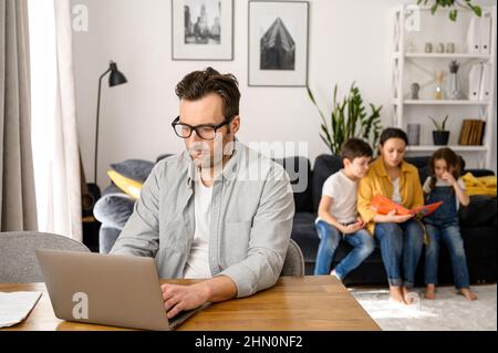 Focused middle-aged father using laptop for freelance work sitting at the desk while mother babysitting with two children, reading book on the background. Dad working on the distance in home office Stock Photo