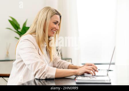 Portrait of young caucasian woman sitting at the desk with laptop, typing an email, messaging to the colleague, chatting with a pleasant smile Stock Photo