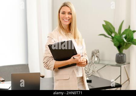 Successful confident young female entrepreneur businesswoman in casual clothes standing with folder at her hands against window and looking at camera, female office employee, real estate agent Stock Photo