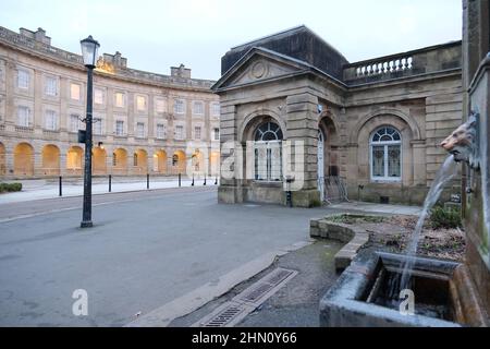 Drinkable Buxton spring water emanating from lion's head fountain at St Ann's Well with Buxton Crescent hotel and Pump Room in the background Stock Photo