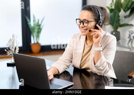 Middle-aged woman wearing wireless headset using a laptop computer for communication with customers or colleagues sitting at the desk in contemporary office. Female support worker talks online Stock Photo