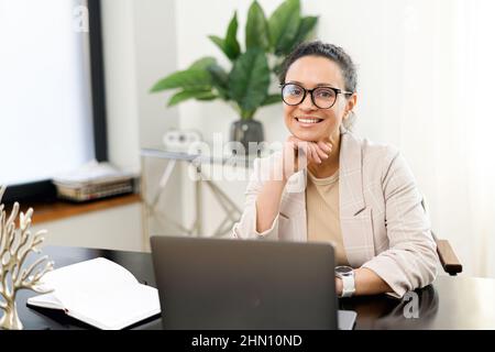 Confident middle-aged woman work with a laptop at the modern office, female employee in smart casual wear looks at the camera and smiles, typing messages, answering email, front view Stock Photo
