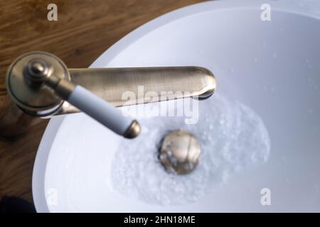 Sink basin faucet, bathroom interior detail. Open bronze tap with running water on white washbasin, close up view from above Stock Photo