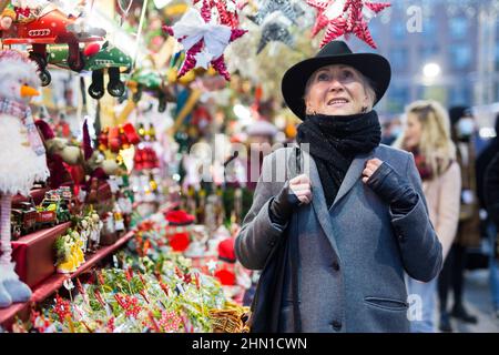 Positive elderly woman in festive mood walking on street Christmas fair Stock Photo