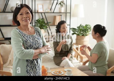Portrait of cheerful elegant senior woman standing in living room, her friends drinking tea and chatting in background Stock Photo