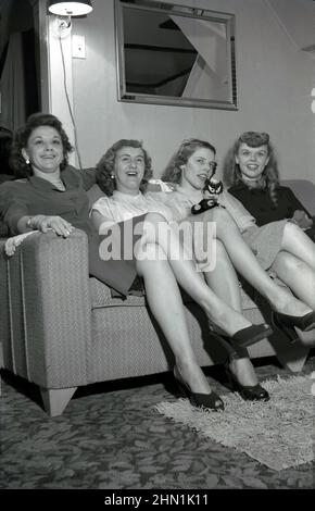 1950s, historical, inside a room, at a party, four ladies, two mothers and their daughers sitting beside each other on a sofa, smile for the camera showing some their shapely legs, USA. Stock Photo