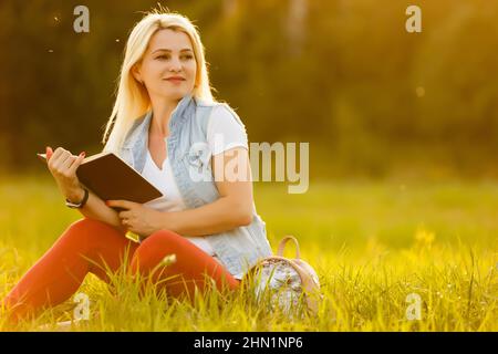 against background of green meadow grass sat young, charming girl, student reads book Stock Photo