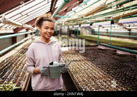 Happy young African-American farmer with watering-pot standing in front of camera against large and long tables with seedlings Stock Photo