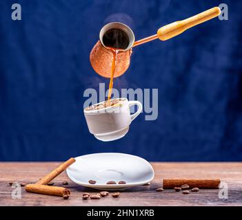 Levitation of pouring coffee from a cezve into a cup with a splash. Cinnamon sticks and coffee beans on the table. Blue background, copy space. Stock Photo