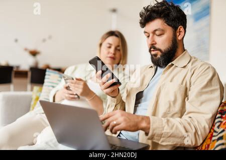 White couple using cellphones and laptop while sitting on couch at home Stock Photo