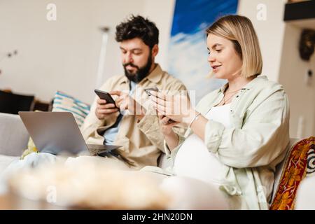Bearded man and pregnant woman using cellphones while sitting on couch at home Stock Photo