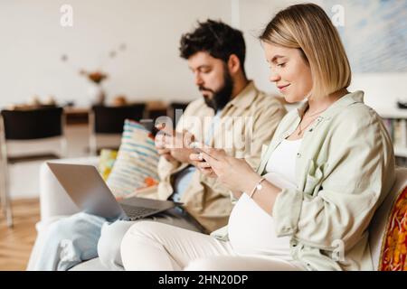 Bearded man and pregnant woman using cellphones while sitting on couch at home Stock Photo