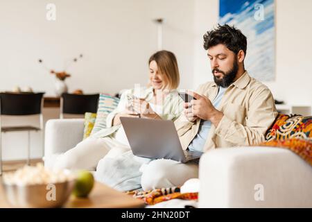White couple using cellphones and laptop while sitting on couch at home Stock Photo