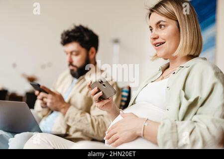 Bearded man and pregnant woman using cellphones while sitting on couch at home Stock Photo