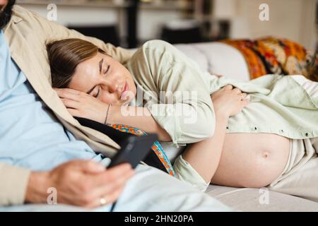 White man using cellphone while his pregnant wife sleeping on couch at home Stock Photo
