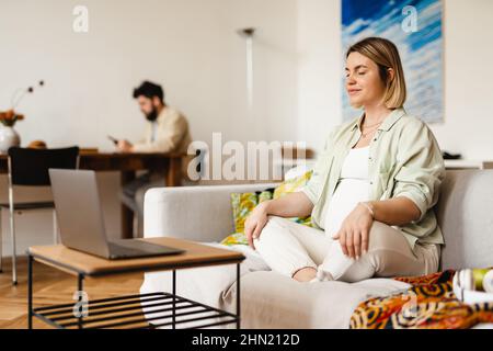 Young pregnant woman meditating and using laptop while sitting on couch at home Stock Photo