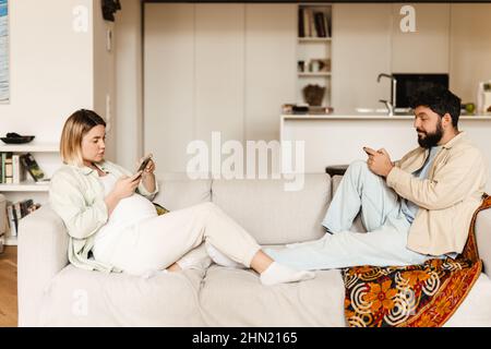 Bearded man and pregnant woman using cellphones while sitting on couch at home Stock Photo